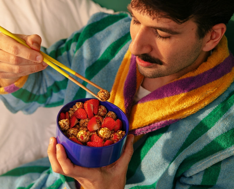 Man in a colorful robe uses yellow chopsticks to eat granola and strawberries from a blue bowl. The setting is cozy, conveying a relaxed morning vibe.