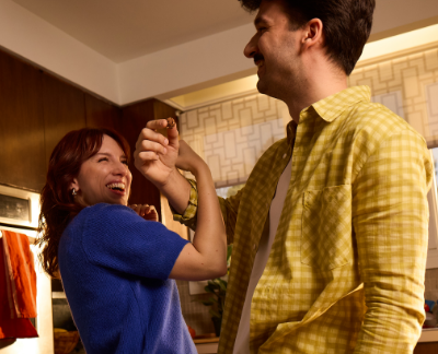 A woman playfully feeds a man in a cozy kitchen. She is smiling and wearing a blue sweater; he has a yellow plaid shirt. The mood is joyful and intimate.