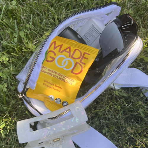 A white purse lies open on green grass, revealing sunglasses, a snack pack labeled "MADE GOOD," and a white hair clip. The scene is relaxed and casual.