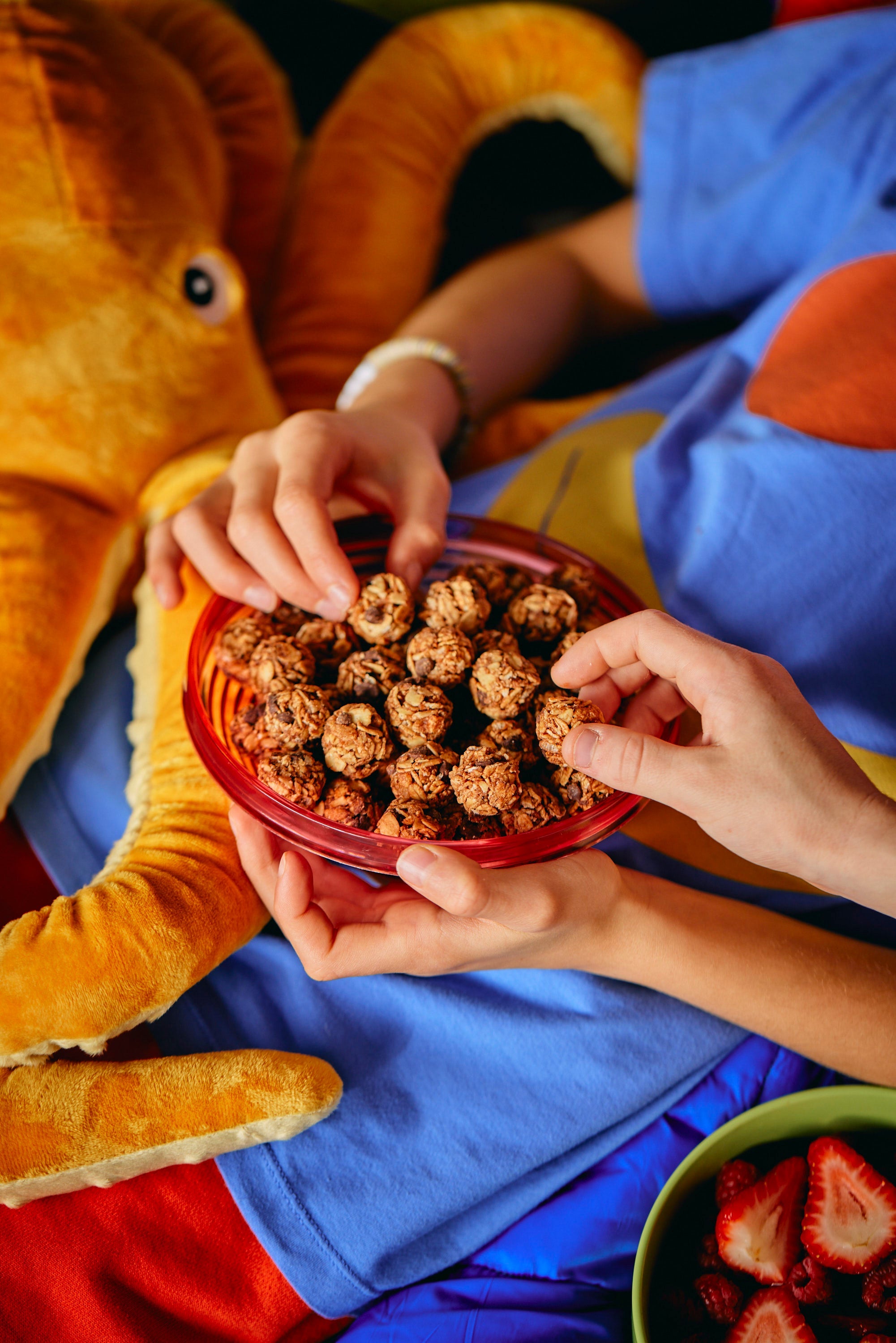 Two hands reach for snacks in a red bowl on a person's lap, who is wearing a blue shirt. A plush orange octopus is in the background. Mood is cozy and playful.