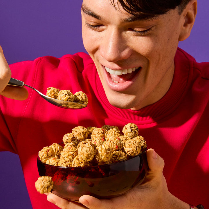 A cheerful young man in a red sweatshirt is holding a bowl filled with MadeGood Granola Bites. He is smiling excitedly as he scoops up a spoonful, ready to take a bite. One snack bite appears to be mid-air, adding a playful touch. The vibrant purple background contrasts with his red outfit, making the scene feel lively and energetic.