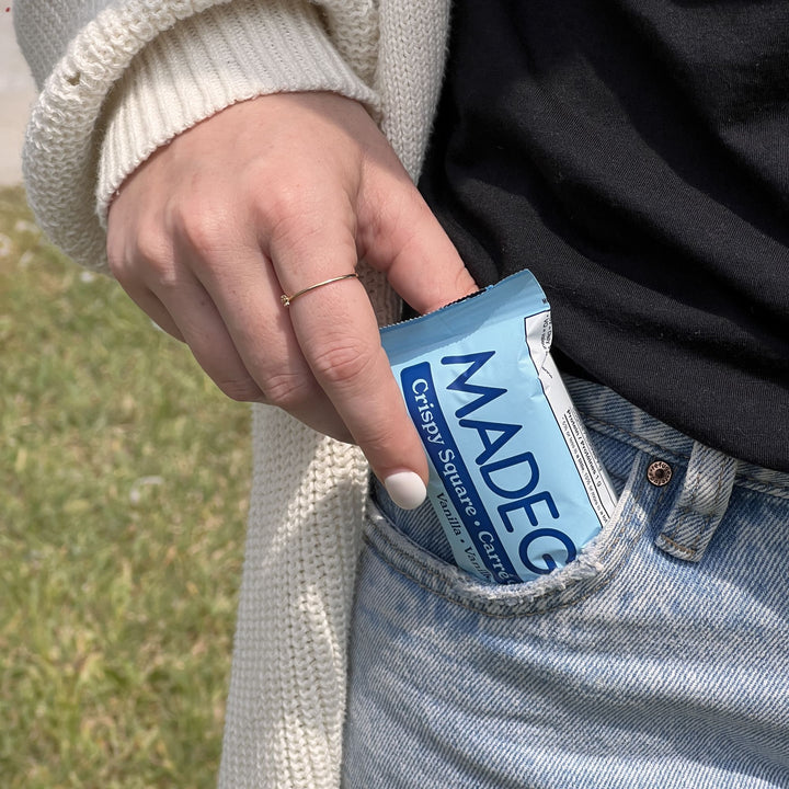 A close-up of a person casually slipping a MadeGood Vanilla Crispy Square into the front pocket of their light-wash denim jeans. They are wearing a black shirt, a cream-colored knit cardigan, and a delicate gold ring on their finger. The background features a grassy outdoor setting.