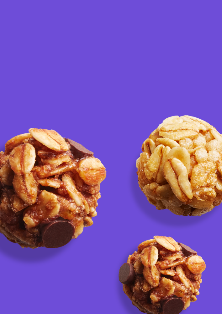 Three round granola bites made of oats, nuts, and chocolate chips against a purple background. Two of the bites have a darker, chocolatey appearance, while the third is lighter in color.