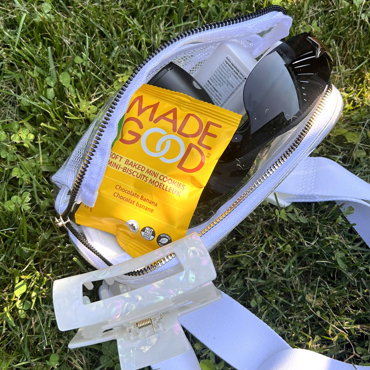 A clear zippered bag rests on lush green grass, partially unzipped to reveal a bright yellow bag of MadeGood Soft Baked Mini Cookies in Chocolate Banana flavor. Also inside the bag are a pair of black sunglasses, a white bottle, and a pearlescent hair claw clip clipped onto the bag’s strap, reflecting the sunlight.