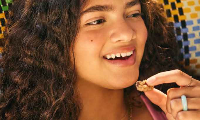 A young person with curly hair smiles, holding a small snack in their hand. The background is a colorful mosaic, creating a joyful and lively atmosphere.