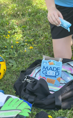 A blue box of MadeGood Vanilla Crispy Squares sits partially open in a black sports bag with a colorful striped interior. Nearby, a yellow soccer ball and a pair of green goalkeeper gloves rest on the grass, while a person in athletic clothing reaches for a snack bar. The setting is outdoors with green grass and small yellow flowers.