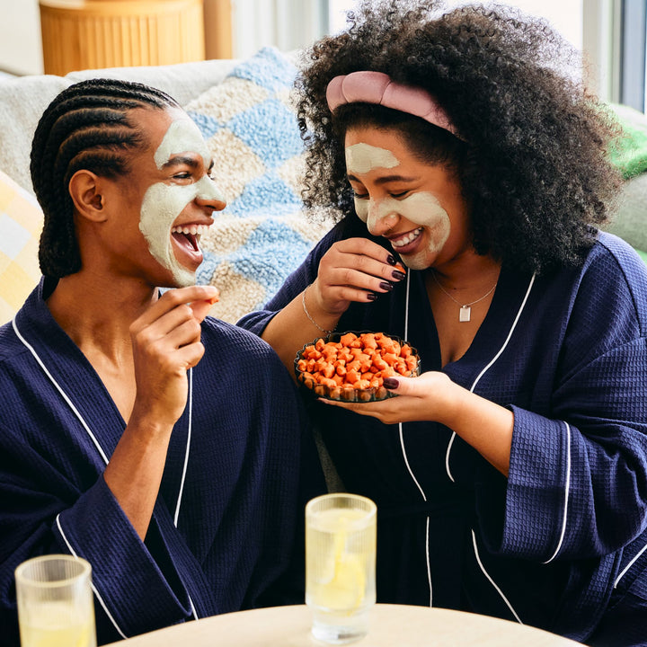 Two friends wearing matching navy blue robes and green facial masks are sitting on a couch, laughing and snacking. One person holds a small bowl filled with MadeGood Star Puffed Crackers, while the other reaches for a piece. They are relaxed, enjoying a cozy self-care moment with drinks on the table.