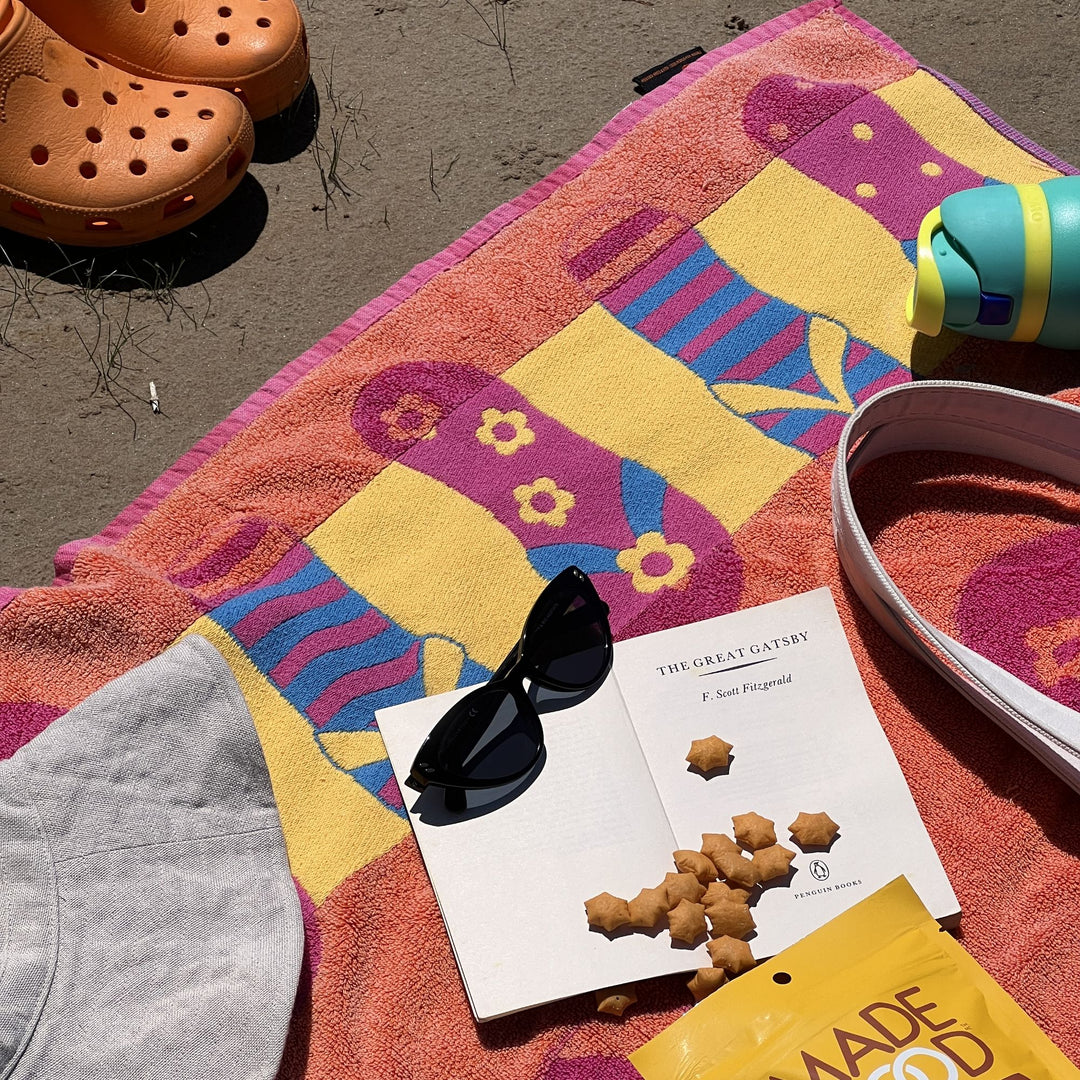 A colorful beach towel with a bold sock pattern is spread on the sand, surrounded by summer essentials. A pair of orange Crocs, a turquoise water bottle, and a gray sun hat are nearby. On the towel, an open copy of The Great Gatsby rests beside a pair of black sunglasses, with star-shaped MadeGood crackers scattered on the pages.