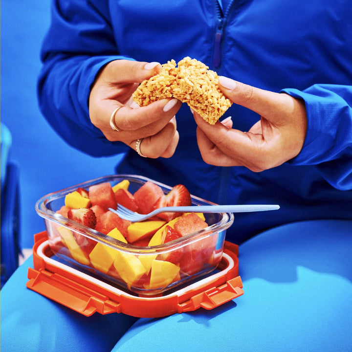 A person in a blue tracksuit breaks apart a crispy square treat while holding a glass container filled with fresh fruit, including watermelon, mango, and strawberries.