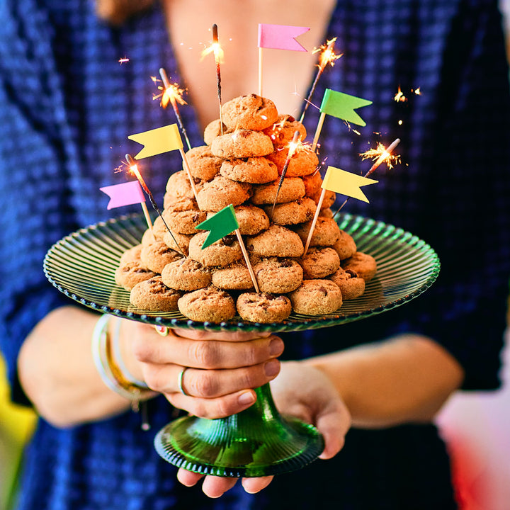 A person in a blue outfit holds a green glass cake stand with a festive cookie tower, decorated with colorful flags and sparklers for a celebratory touch.