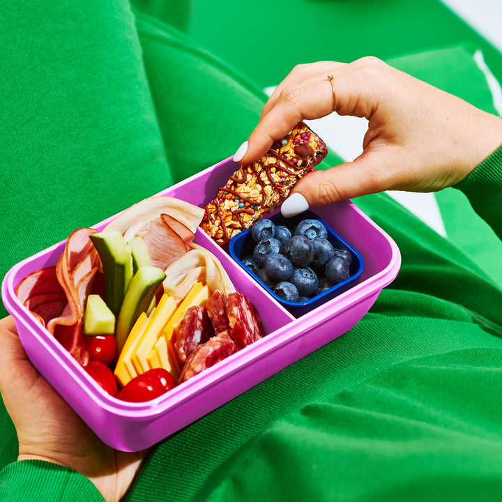 A person in a green outfit holds a purple bento box filled with fresh ingredients, including cheese, meats, cucumbers, cherry tomatoes, and blueberries. They pick up a chocolate-drizzled granola bar, adding a sweet crunch to the balanced snack.