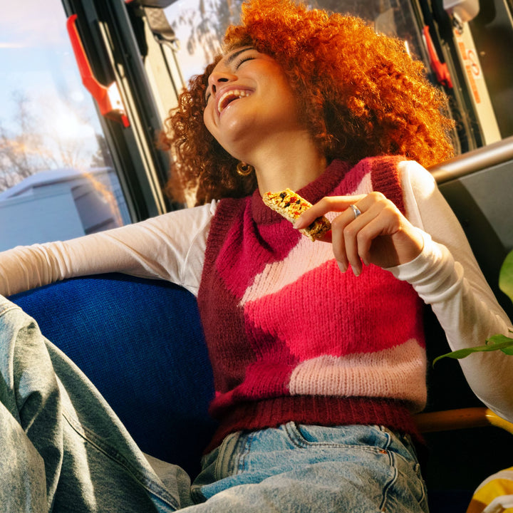 A young woman with curly red hair leans back in a blue seat on a bus, laughing with her head tilted back. She wears a white long-sleeve shirt under a pink and burgundy patterned knit vest, along with light-wash jeans. Sunlight streams through the window, casting a warm glow on her face. She holds a granola bar in one hand, while the other rests casually on the armrest, enjoying a relaxed and joyful moment.