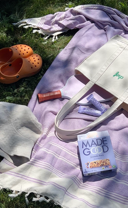 A picnic scene on a purple patterned blanket laid on green grass. Items include orange clogs, a beige bucket hat, a white tote bag with 'Jeep' embroidered in green, a lavender pickleball paddle, sunscreen, and a box of MadeGood Mixed Berry Granola Bars.