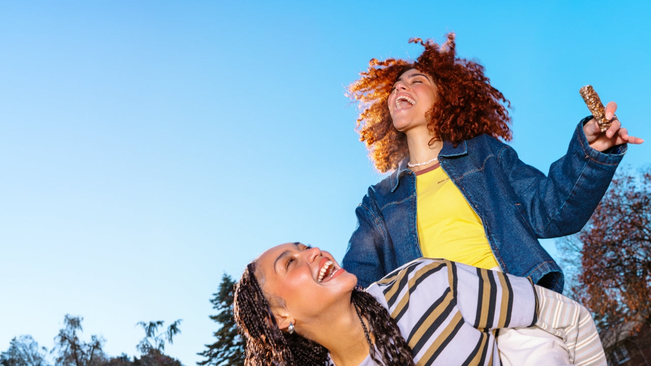 Two people outdoors against a blue sky, laughing joyfully. One in a striped shirt leans forward, while the other with curly hair raises a snack bar.