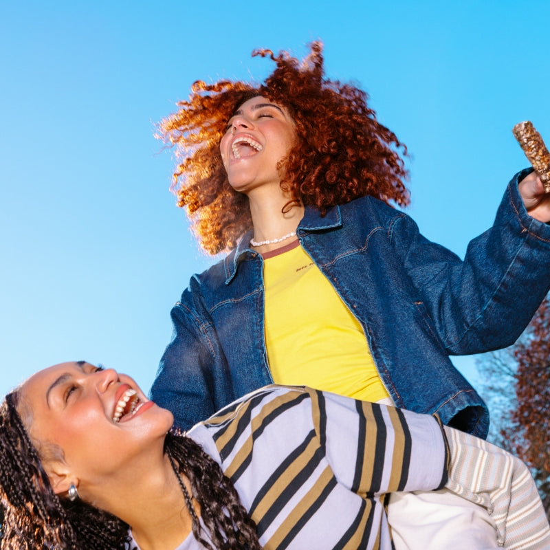 Two people outdoors against a blue sky, laughing joyfully. One in a striped shirt leans forward, while the other with curly hair raises a snack bar.