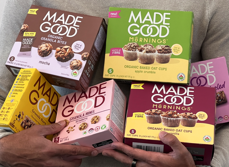 Two hands hold a variety of Made Good snack boxes, including granola bites and oat cups. The packaging is colorful, conveying a wholesome and nutritious vibe.