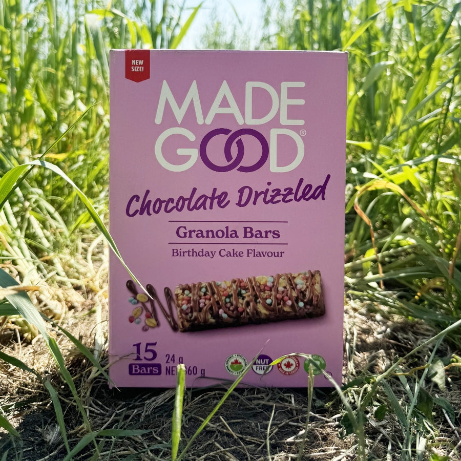 Box of "Made Good" chocolate drizzled granola bars with birthday cake flavor in a lush field. Vibrant greenery surrounds the purple packaging.