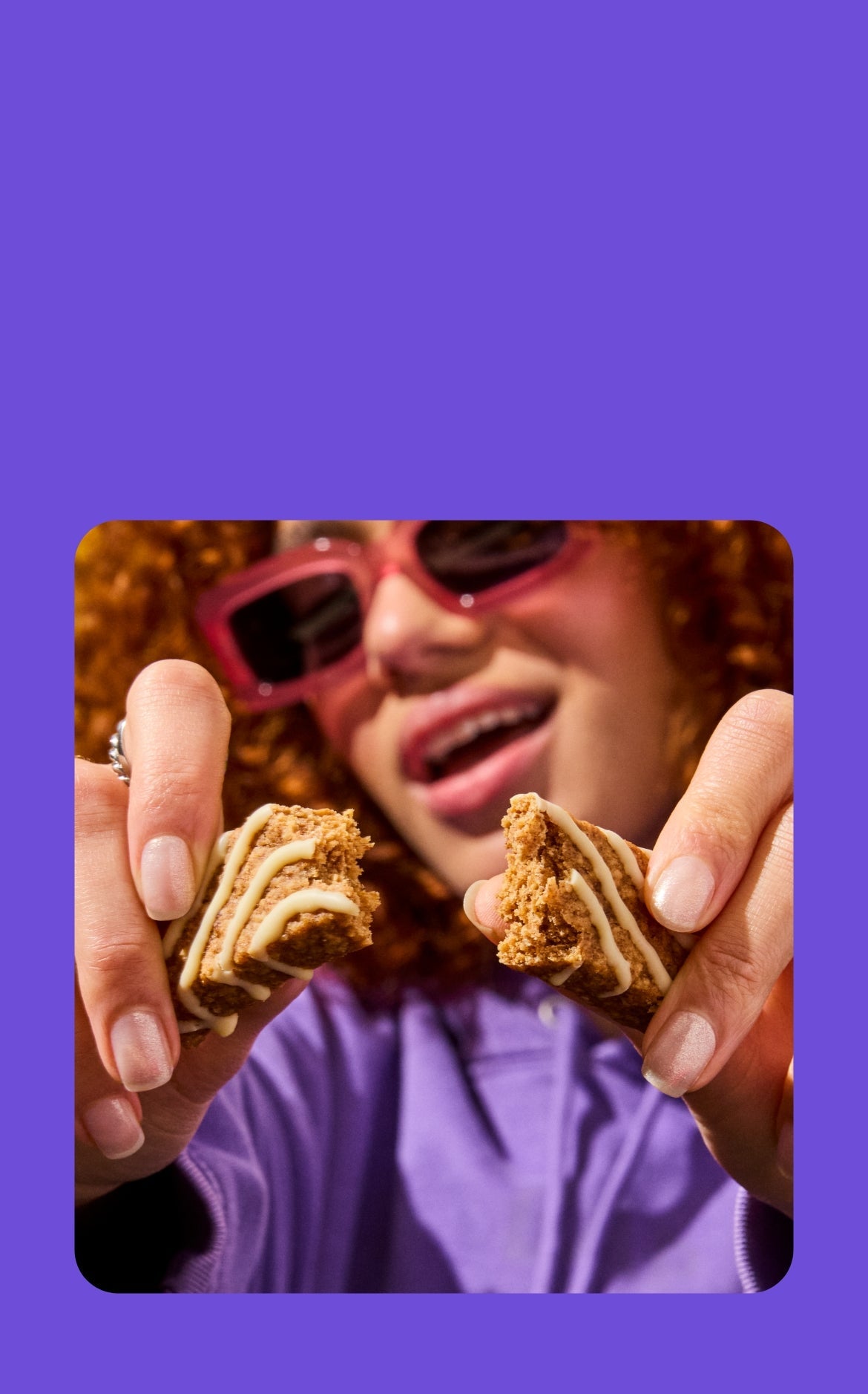 A young woman with curly red hair and pink sunglasses smiles as she holds and breaks apart a Mornings soft baked breakfast oat bar. She is wearing a purple sweater, and the bar’s soft, crumbly texture is visible, with icing drizzled in a zigzag pattern.