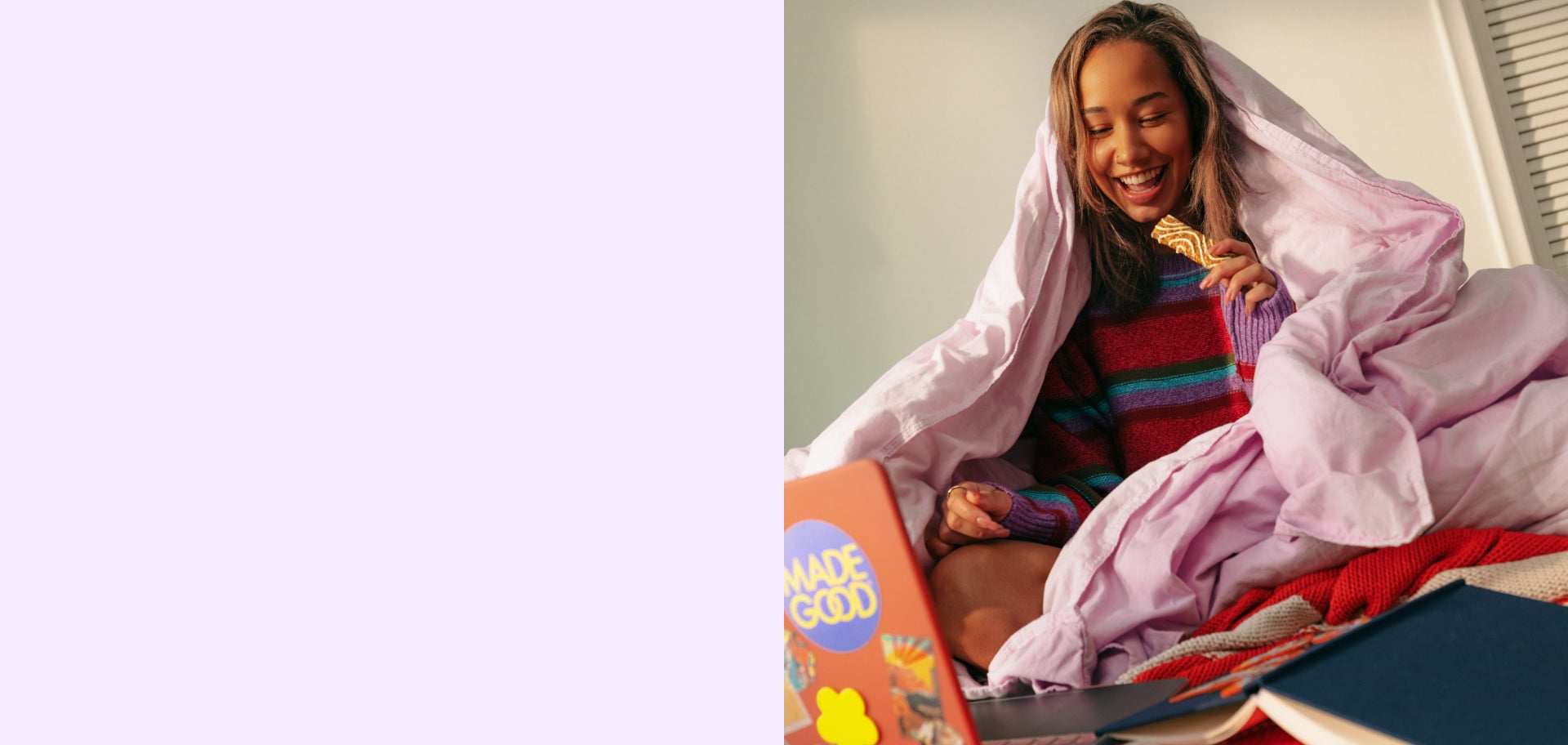 A girl with light brown hairs site on her bed with a blanket over her head while she happily eats a morning bar.