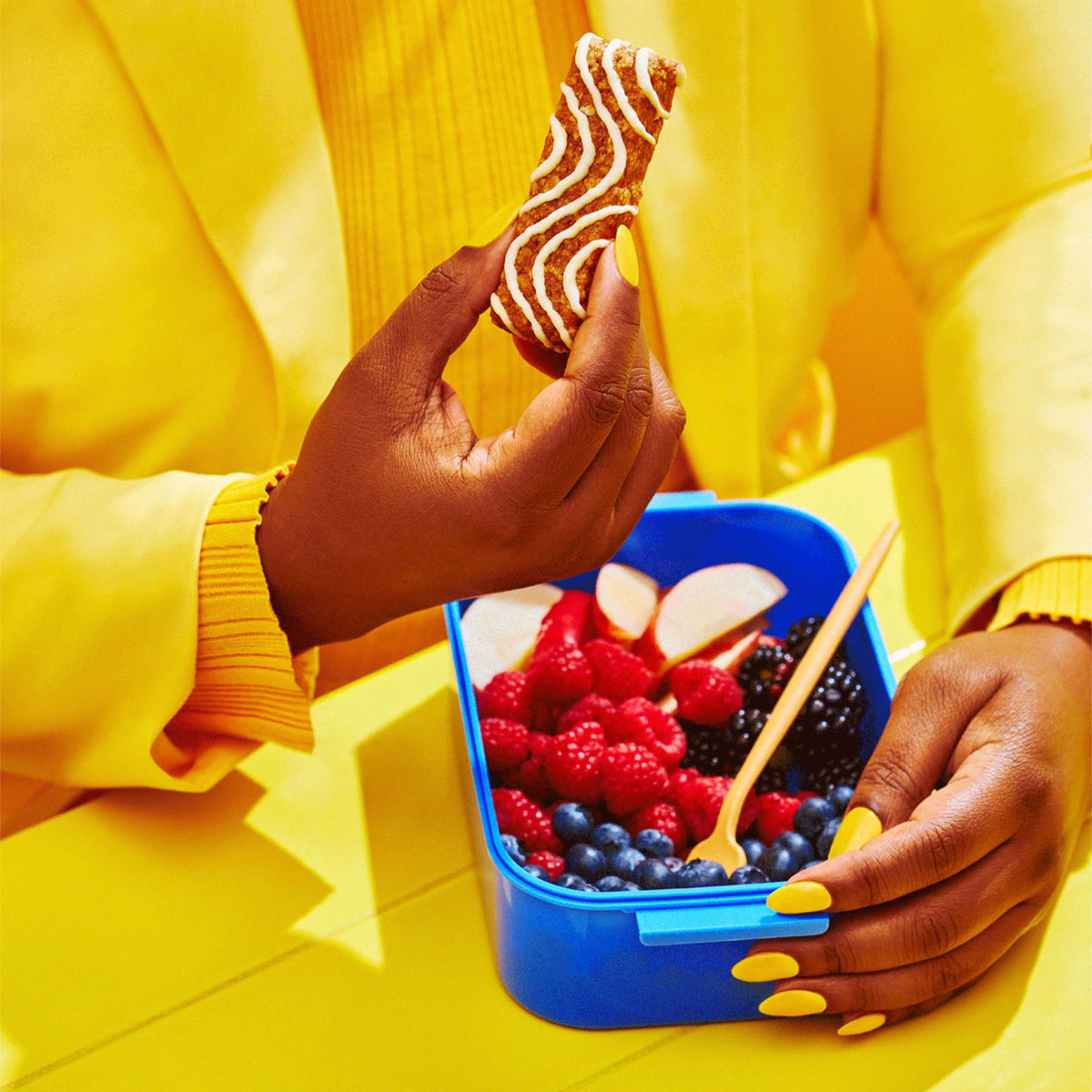 A person wearing a yellow sweater and matching nail polish holds a Mornings soft baked oat bar in one hand while the other rests on a blue lunchbox filled with fresh raspberries, blueberries, blackberries, and sliced apples. The background is also bright yellow, creating a bold, monochromatic look.