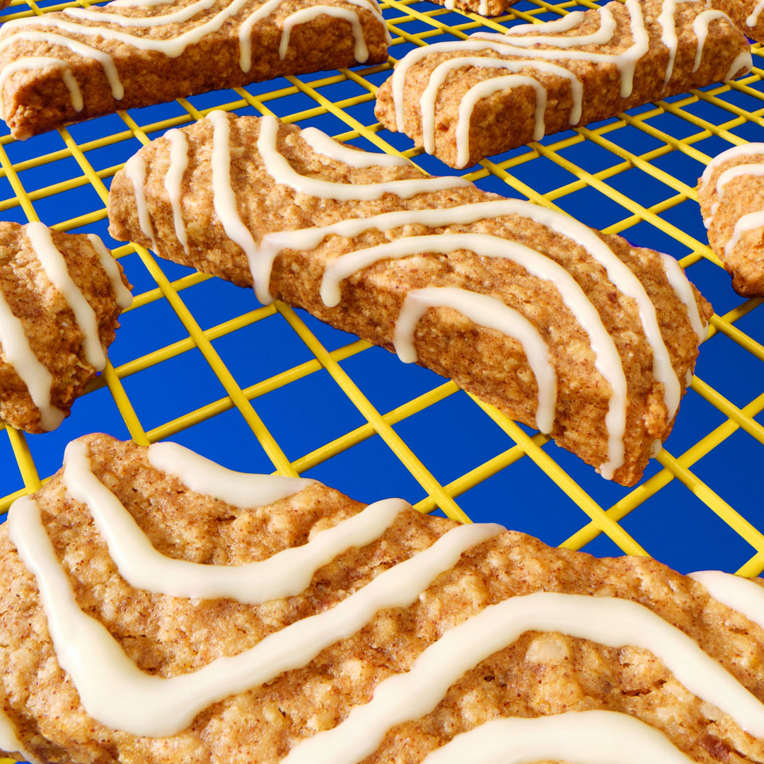 A close-up of Mornings soft baked oat bars with a drizzle of white icing, cooling on a bright yellow wire rack against a bold blue background. The bars have a textured surface, and the icing is applied in wavy lines.