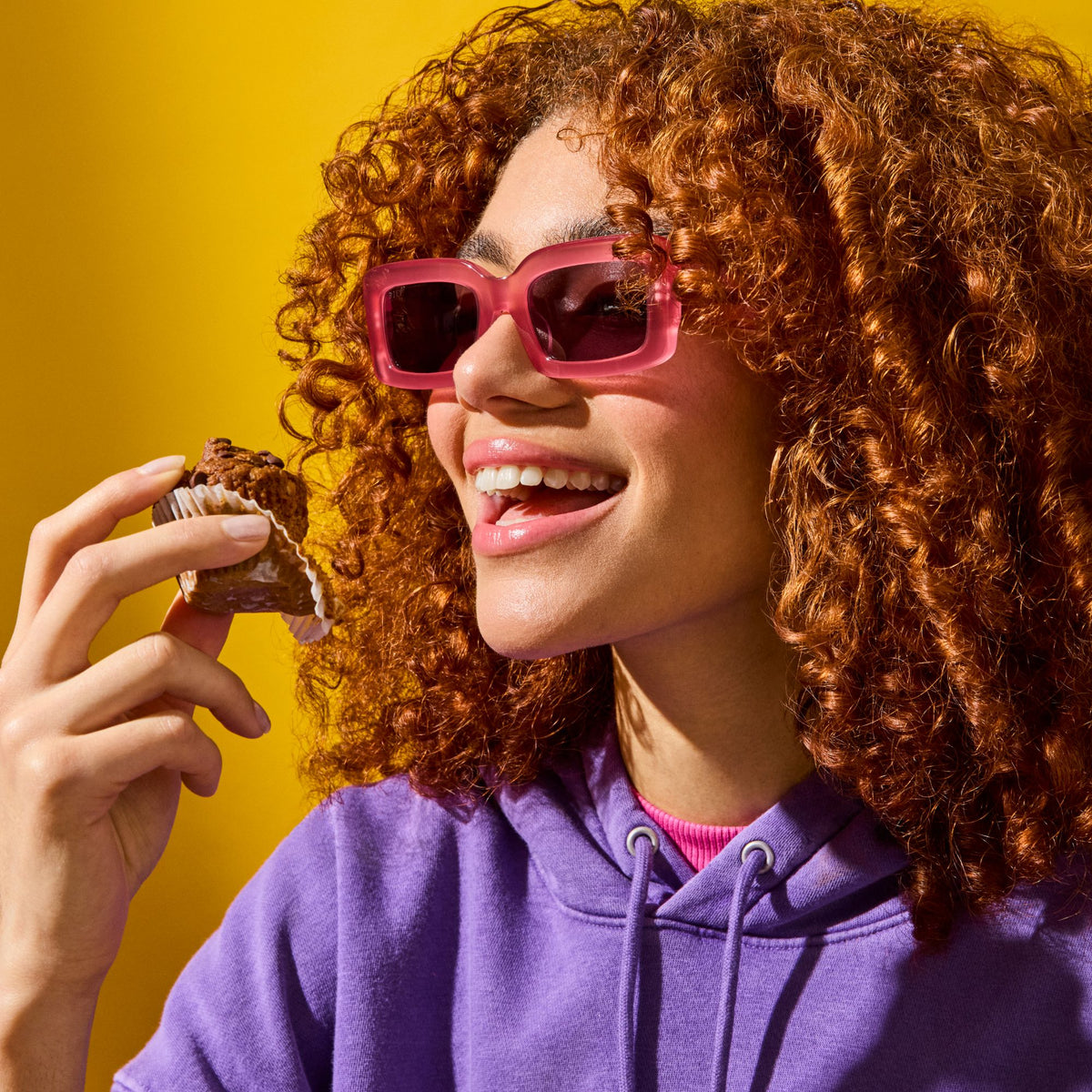 A young woman with voluminous curly red hair and pink sunglasses smiles as she holds a Mornings Oat Cup in a paper wrapper. She is wearing a purple hoodie and a pink turtleneck, with a bright yellow background adding vibrancy to the image. The scene exudes joy and indulgence.