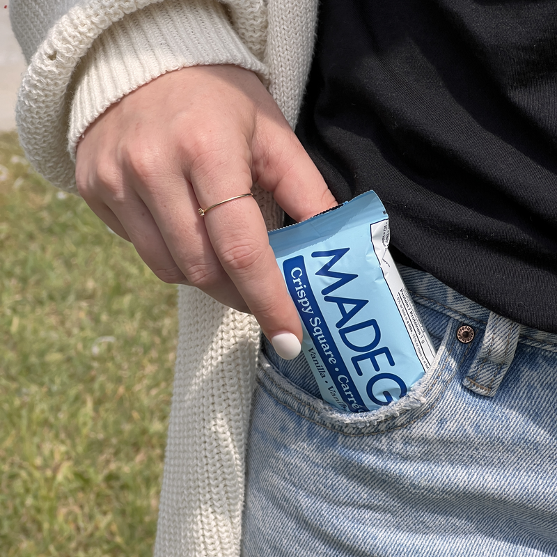 A hand placing a blue-packaged snack into a pocket of light blue jeans. The person wears a cream cardigan, with green grass in the background.