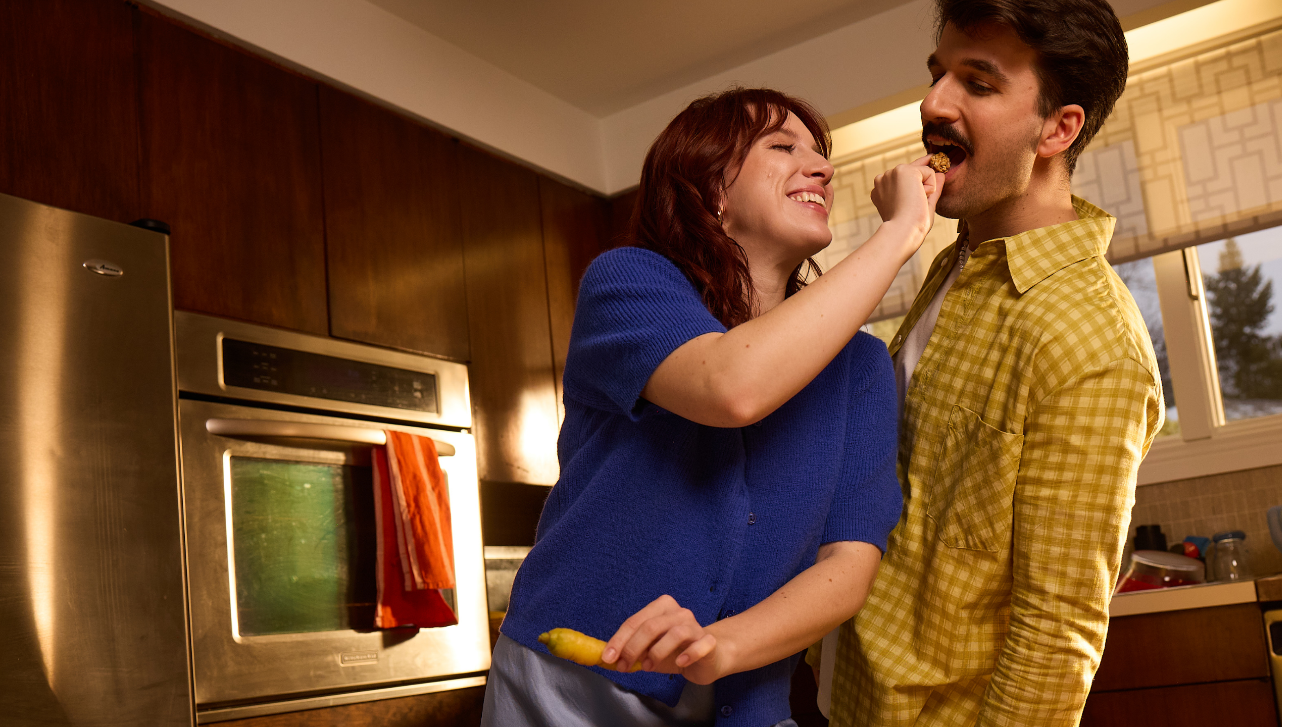 A woman playfully feeds a man in a cozy kitchen. She is smiling and wearing a blue sweater; he has a yellow plaid shirt. The mood is joyful and intimate.