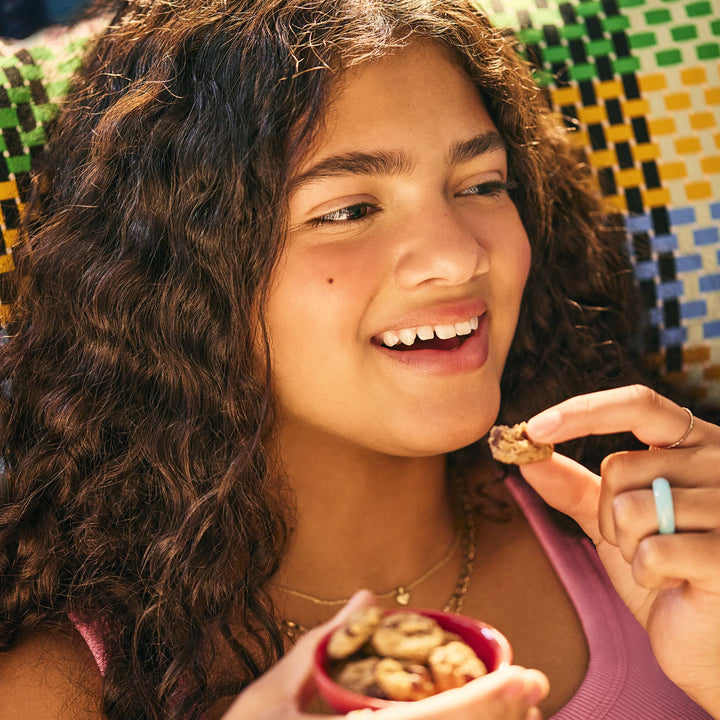 A young woman with curly brown hair reclines on a colorful woven chair, smiling as she enjoys a bite-sized soft-baked cookie. She holds a small red bowl filled with more cookies in one hand, while the other lifts a piece to her mouth. She wears a pink tank top, layered gold necklaces, and a turquoise ring, basking in the warm sunlight.