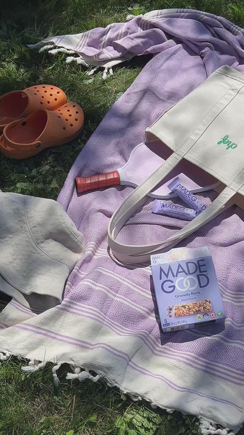 A picnic scene on a purple patterned blanket laid on green grass. Items include orange clogs, a beige bucket hat, a white tote bag with 'Jeep' embroidered in green, a lavender pickleball paddle, sunscreen, and a box of MadeGood Mixed Berry Granola Bars.